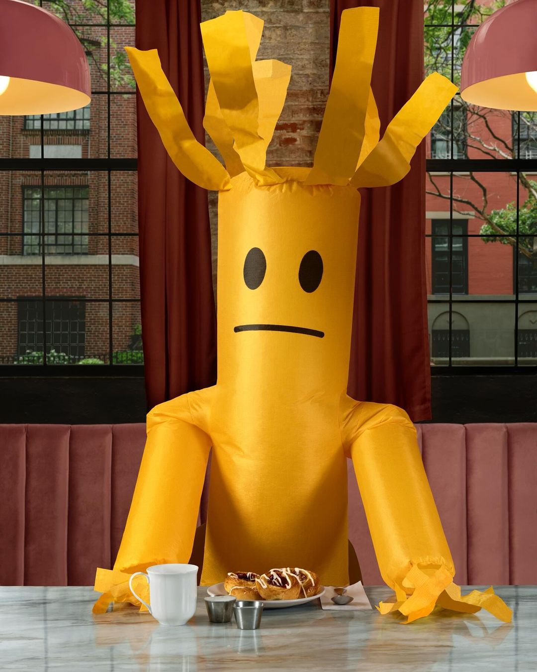 Inflatable tube man with sad face sits at marble table inside a restaurant with pastries, a mug, and condiments.